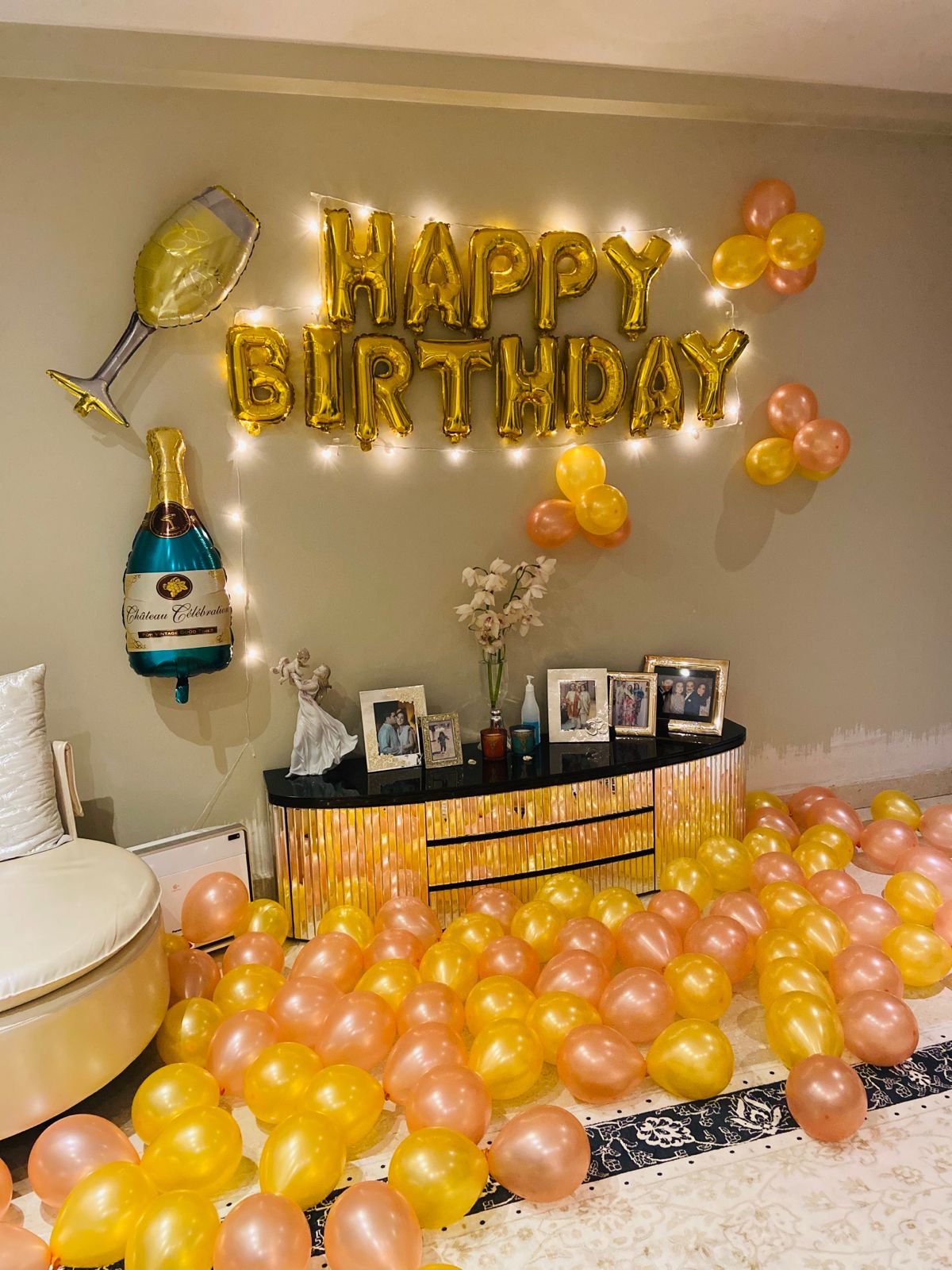 Premium golden and peach birthday decoration with Happy Birthday foil letters, warm fairy lights, golden and peach balloons, champagne and wine glass foil balloons, balloon clusters and floor balloon decoration for an elegant birthday celebration at home in Delhi NCR.